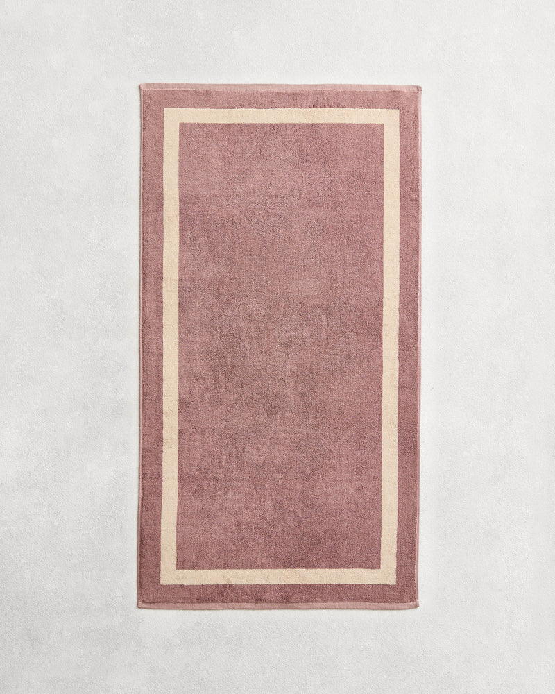 Flat cotton terry towel on a grey background in a lavender colour with a creme border.