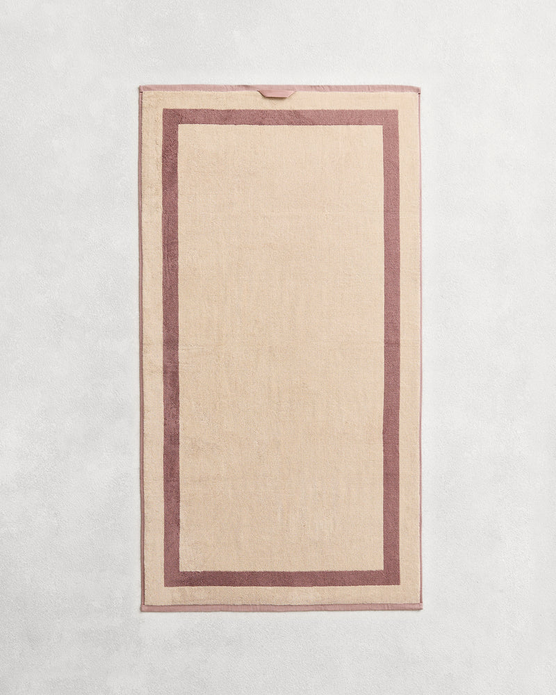 Flat cotton terry towel on a grey background in a creme colour with a lavender border.