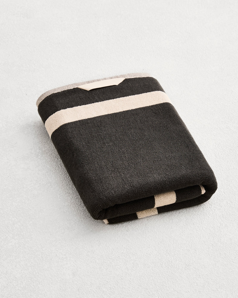 A folded cotton terry bath towel on a grey background, in a charcoal colourway with an oatmeal border. 
