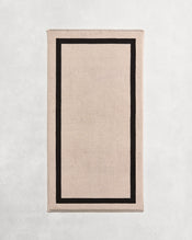 Flat cotton terry bath towel on a grey background in an oatmeal colour with a charcoal border. - view