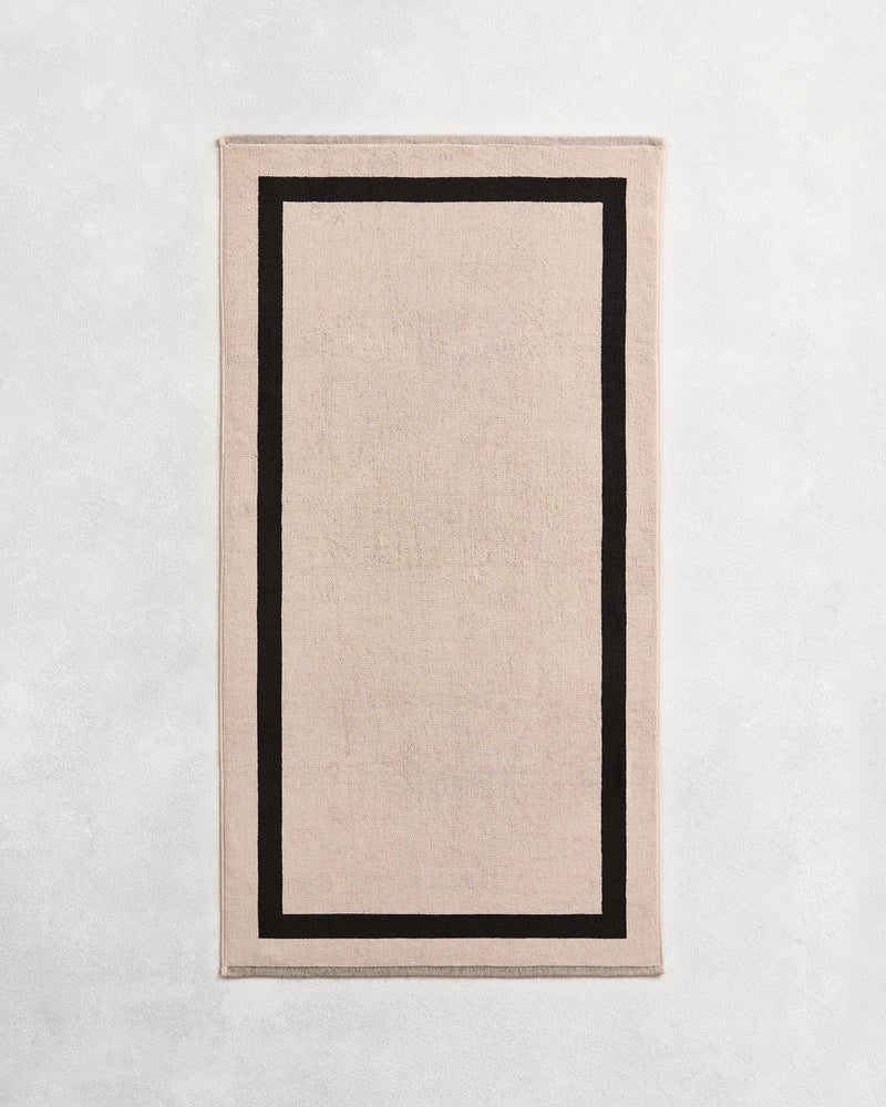 Flat cotton terry bath towel on a grey background in an oatmeal colour with a charcoal border.