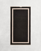 Flat cotton terry bath towel on a grey background in a charcoal colour with an oatmeal border. - view