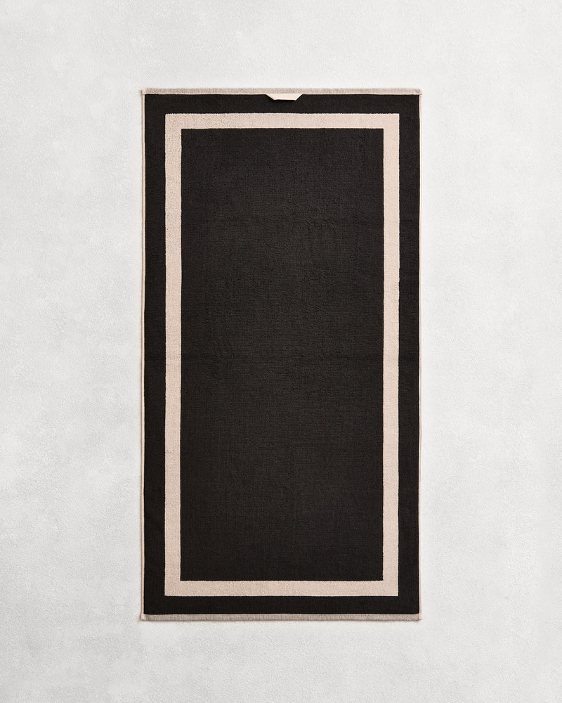 Flat cotton terry bath towel on a grey background in a charcoal colour with an oatmeal border.