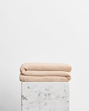 A folded wool cotton blanket in the shade latte, a cream colour, stacked on top of a white marble pedestal against a plain white wall. - view