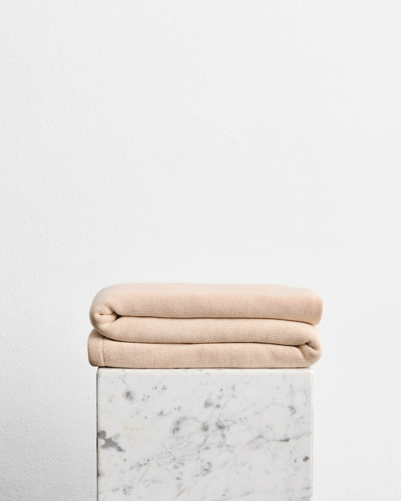 A folded wool cotton blanket in the shade latte, a cream colour, stacked on top of a white marble pedestal against a plain white wall.