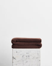 A folded dark brown wool cotton blanket in the shade mocha, stacked on top of a white marble pedestal against a plain white wall. - view