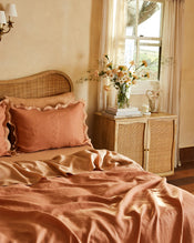 A cozy bedroom features a rattan headboard, peach bedding, Bed Threads Hazelnut & Terracotta 100% French Flax Linen Scalloped Pillowcases, a wooden nightstand with books and flowers, and sunlight filtering through sheer curtains. - view