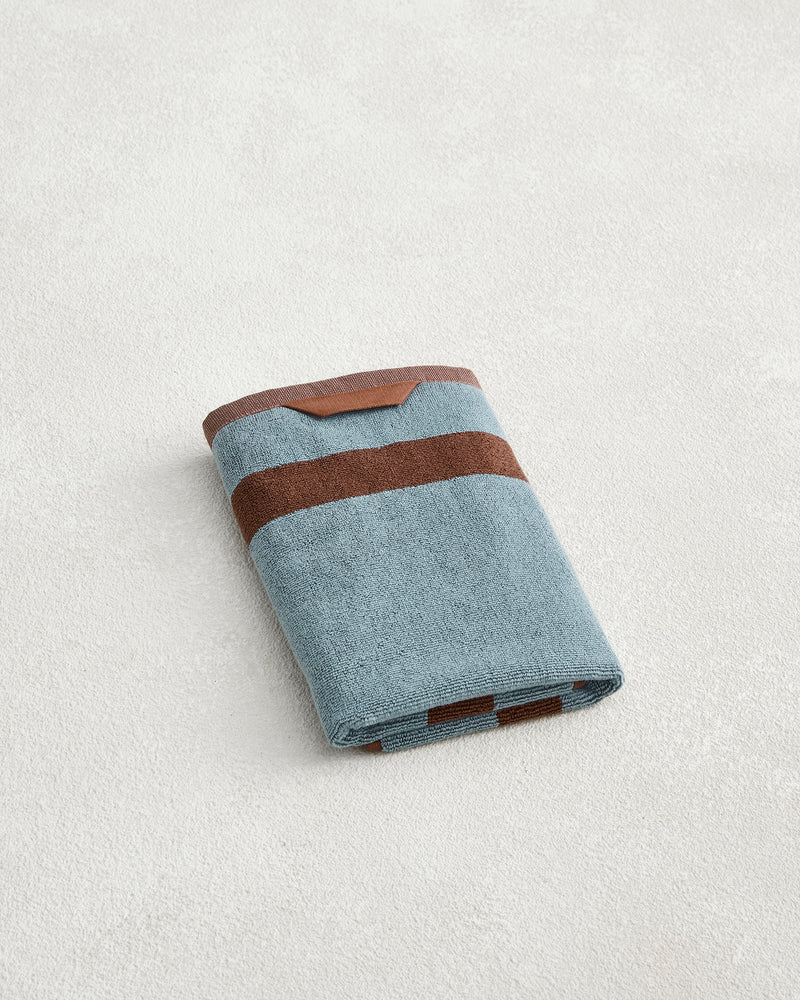 A neatly folded Bed Threads Cacao & Lagoon Two-Toned 100% Cotton Terry Hand Towel with two horizontal brown stripes rests on a light textured surface.