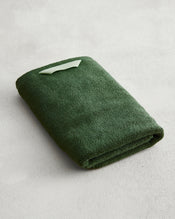 The Bed Threads Moss 100% Cotton Terry Hand Towel, in dark green with a light green label, is neatly folded on a light grey textured surface. Crafted from plush 650GSM cotton, it offers softness and exceptional absorbency. - view