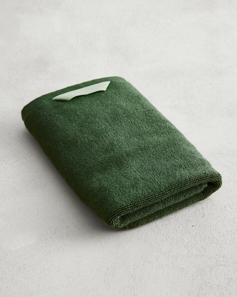 The Bed Threads Moss 100% Cotton Terry Hand Towel, in dark green with a light green label, is neatly folded on a light grey textured surface. Crafted from plush 650GSM cotton, it offers softness and exceptional absorbency.