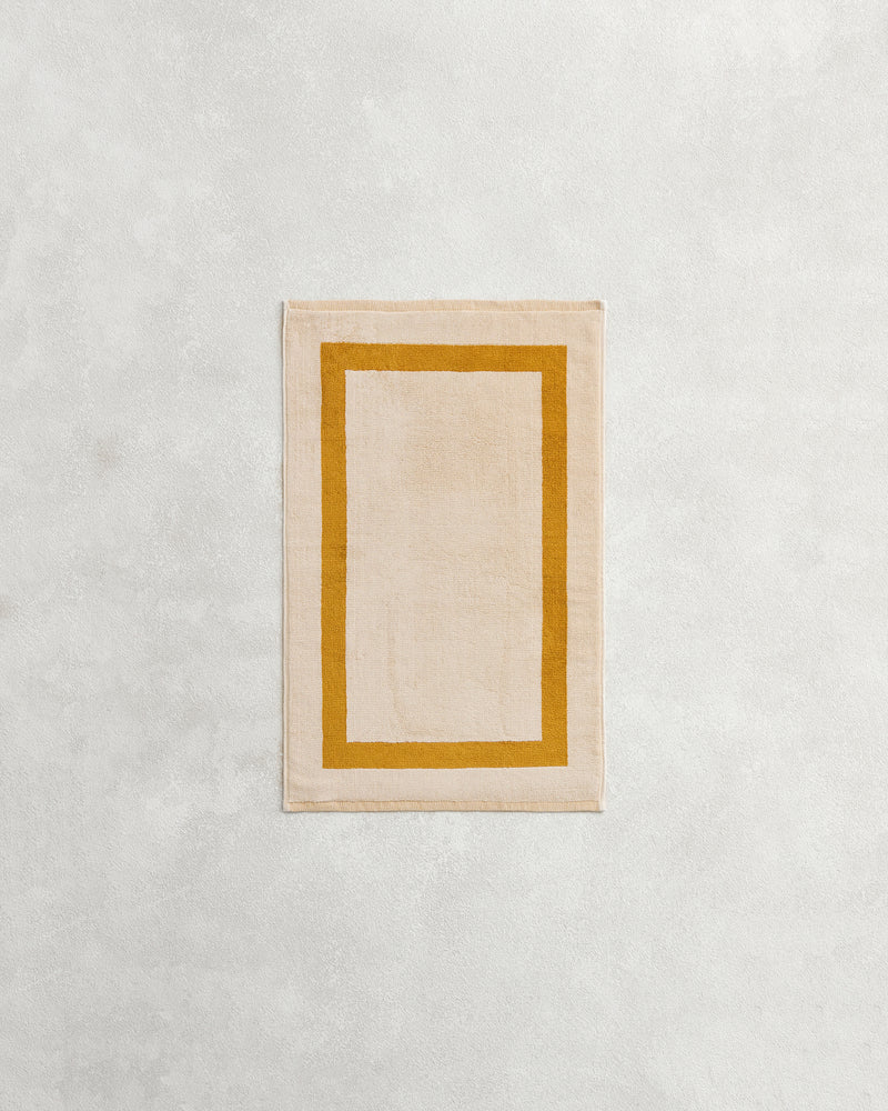 A rectangular beige rug with a mustard border rests on a light grey surface, reflecting the plush 650GSM softness and two-toned style of Bed Threads' Crème & Turmeric 100% Cotton Terry Hand Towel.