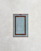 The Cacao & Lagoon Two-Toned 100% Cotton Terry Hand Towel from Bed Threads features a rectangular blue design with a dark brown border and matching inner outline, displayed on a light textured background. - view