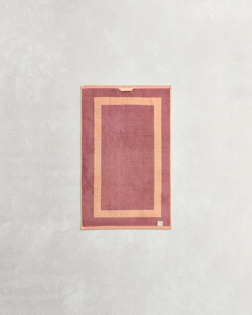 The Bed Threads Terracotta & Pink Clay Two-Toned 100% Cotton Terry Hand Towel, made from plush 650GSM quick-drying terry, features a lighter pink border, small fabric loop, and tag at the bottom corner.