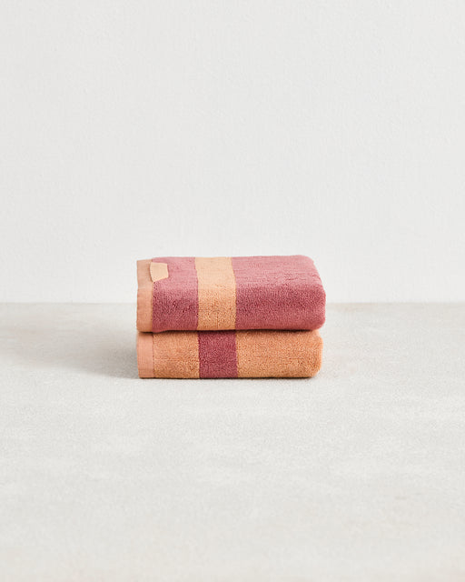 A neatly folded set of two Bed Threads Terracotta & Pink Clay Two-Toned 100% Cotton Terry Hand Towels is stacked on a smooth surface, displaying wide stripes in soft hues against a plain white background.