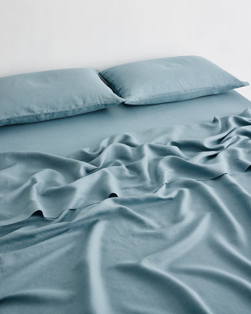 A neatly made bed with Bed Threads' Lagoon 100% French Flax Linen Sheet Set and two matching pillows, the light blue linen slightly rumpled at the center, sits against a plain white wall—ideal for those seeking temperature-regulating bedding.