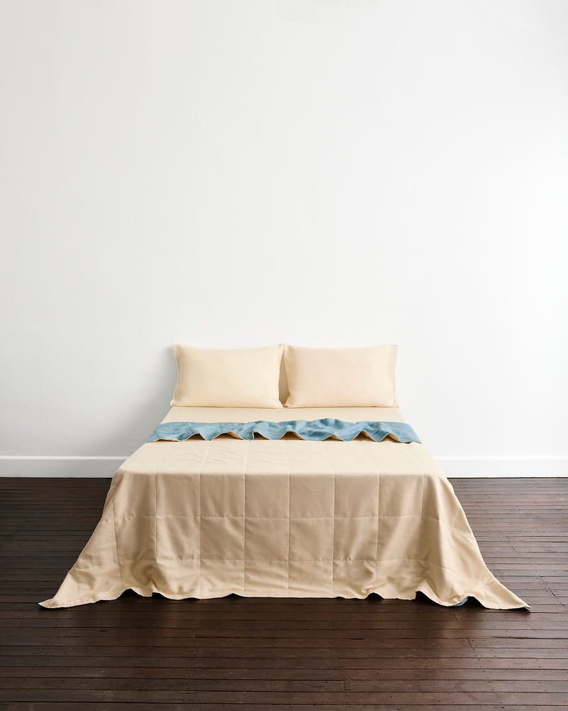A neatly made bed with a cream quilt cover in Lagoon & Creme and matching pillowcases in Creme, set against a plain white wall and dark wood flooring.