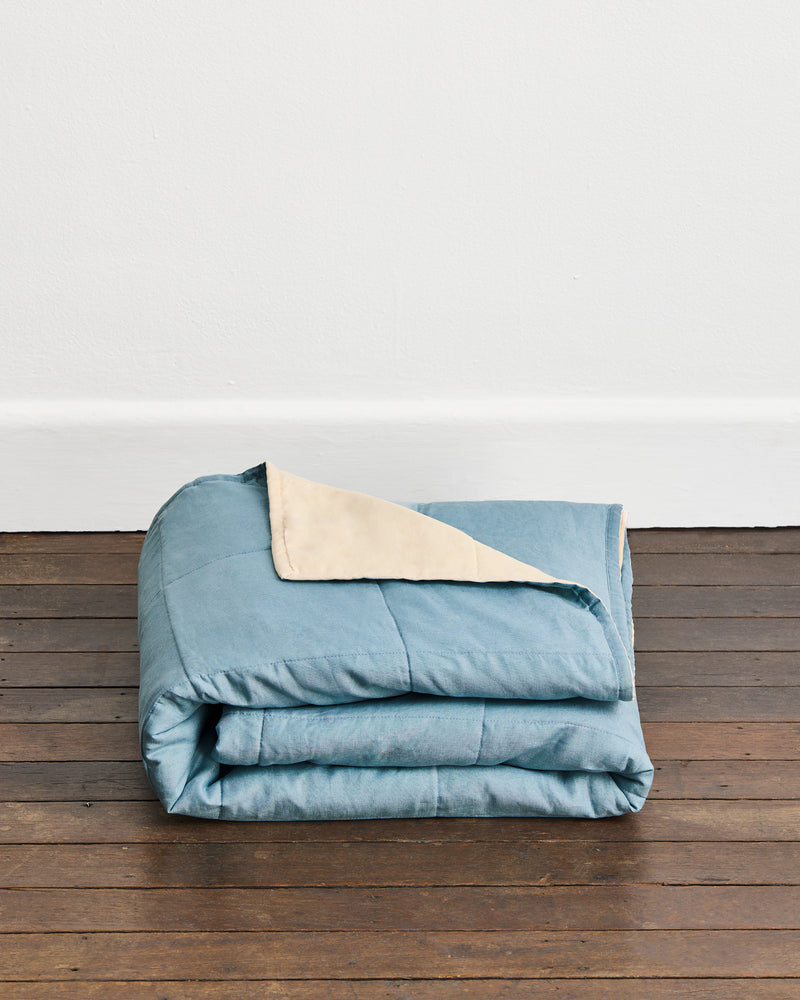 A folded dusty blue quilt cover in Lagoon & Creme with a cream underside, displayed on a dark wooden floor against a white wall.