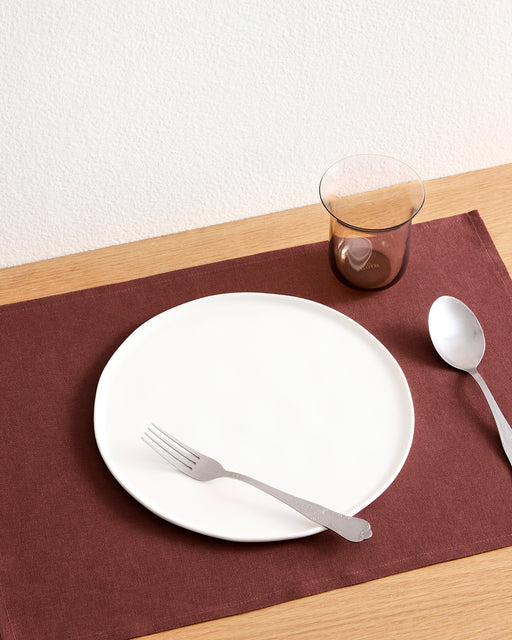 A fork rests on a Bed Threads Cacao 100% French Flax Linen Placemat (Set of Four), beside a metal spoon and a transparent brown cup, all arranged on a light wooden table for elegant protection.