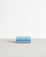 A set of two Sky 100% Cotton Terry Face Washers by Bed Threads, in light blue, sits neatly folded on a light gray surface against a white textured wall—perfect for daily care. - view
