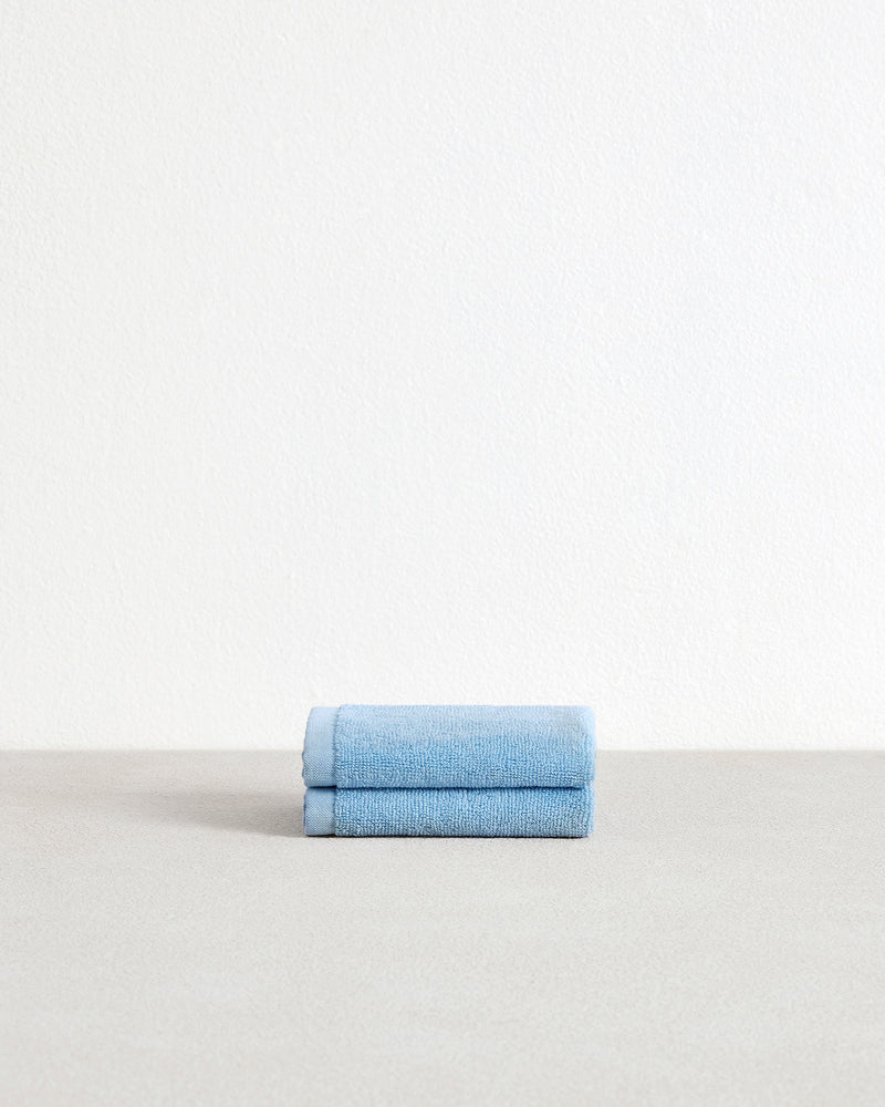 A set of two Sky 100% Cotton Terry Face Washers by Bed Threads, in light blue, sits neatly folded on a light gray surface against a white textured wall—perfect for daily care.