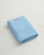 The Bed Threads Sky 100% Cotton Terry Bath Towel with a pink tag is folded on a textured white surface, highlighting its plush softness and premium quality. - view