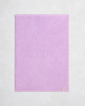 The Bed Threads Violet 100% Cotton Terry Bath Mat, featuring a small fabric tag in the bottom right corner, is neatly arranged and lies flat on a light textured surface. - view