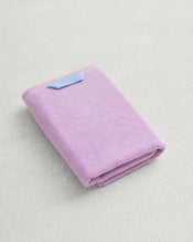 The Bed Threads Violet 100% Cotton Terry Hand Towel, made from plush 650GSM cotton for quick drying, is neatly folded with a blue tag and displayed on a light-colored textured surface. - view