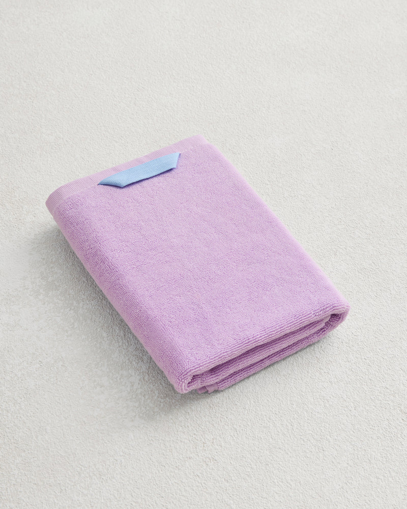 The Bed Threads Violet 100% Cotton Terry Hand Towel, made from plush 650GSM cotton for quick drying, is neatly folded with a blue tag and displayed on a light-colored textured surface.