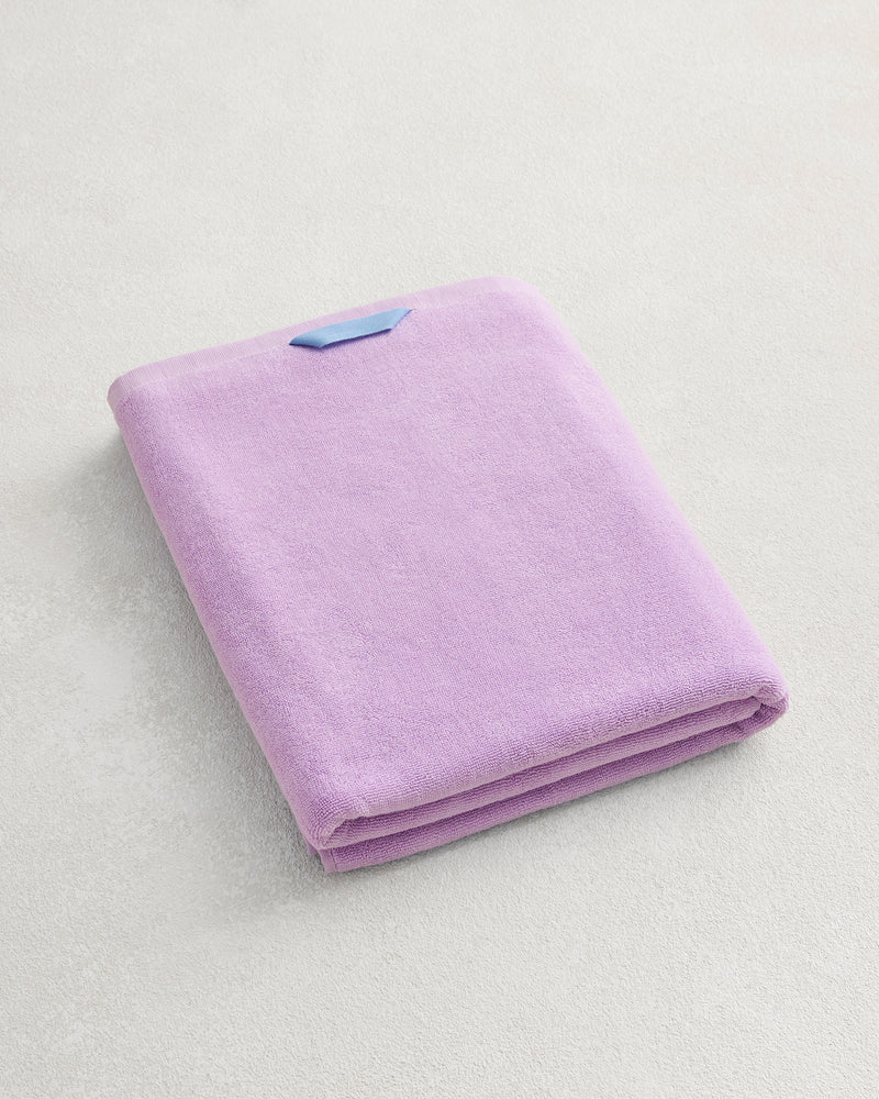 The Bed Threads Violet 100% Cotton Terry Bath Sheet, neatly folded with a blue label, rests on a light textured surface, showcasing the elegance and quality of premium cotton.
