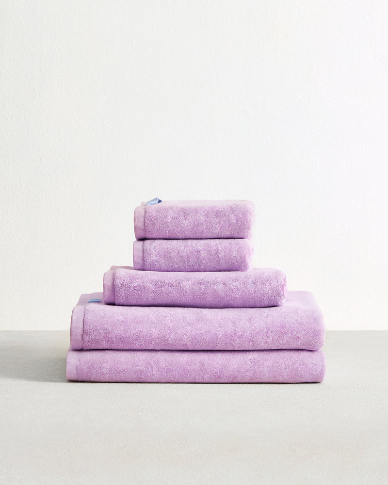 The Bed Threads Violet 100% Cotton Terry Bath Sheet Bundle, featuring five highly absorbent towels in varying sizes, is neatly stacked on a light surface against a plain white background.