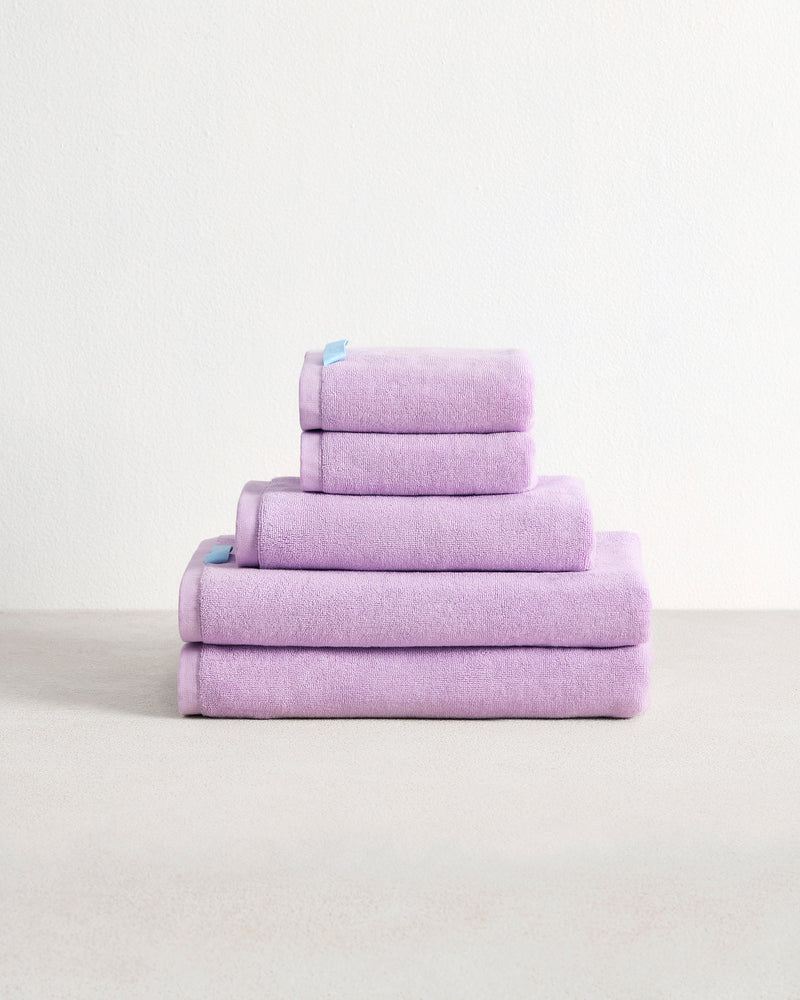 A stack of four Violet 100% Cotton Terry Bath Towels by Bed Threads, in assorted sizes, is neatly folded in a pyramid shape on a light surface with a plain white background.