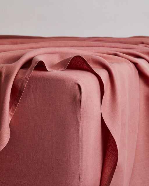 A close-up of a bed corner shows the Bed Threads Pink Clay 100% French Flax Linen Fitted Sheet, softly draped and folded over the edge against a plain, light background.
